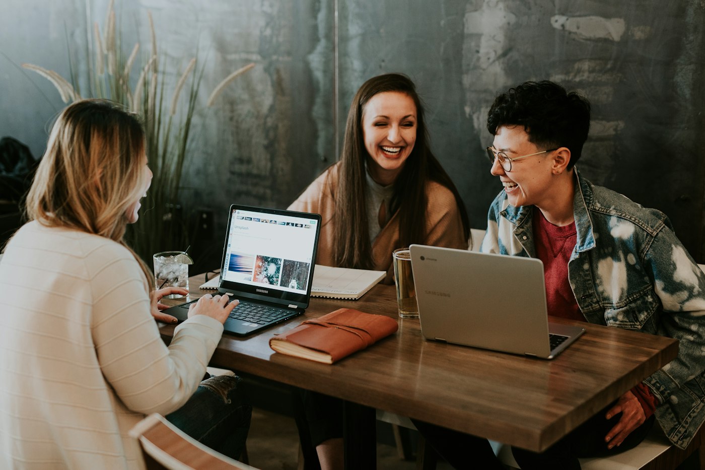 Three people laughing around a wooden table with laptops open